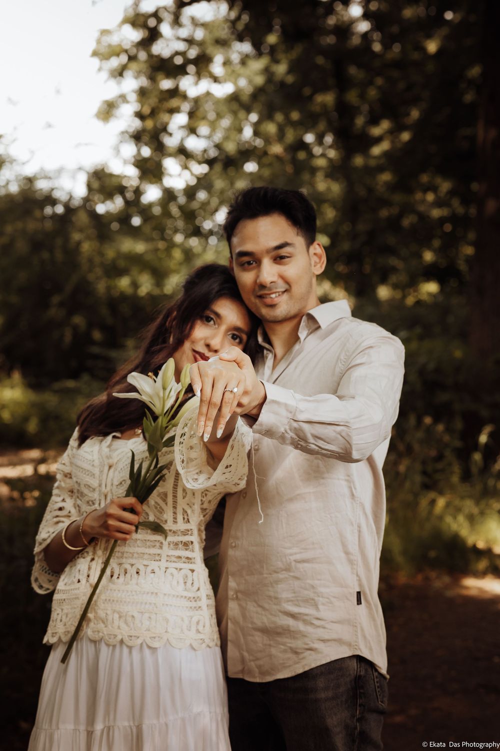 Couple in white outfits showing engagement ring outdoors, woman holding white lily flowers in a romantic forest setting.