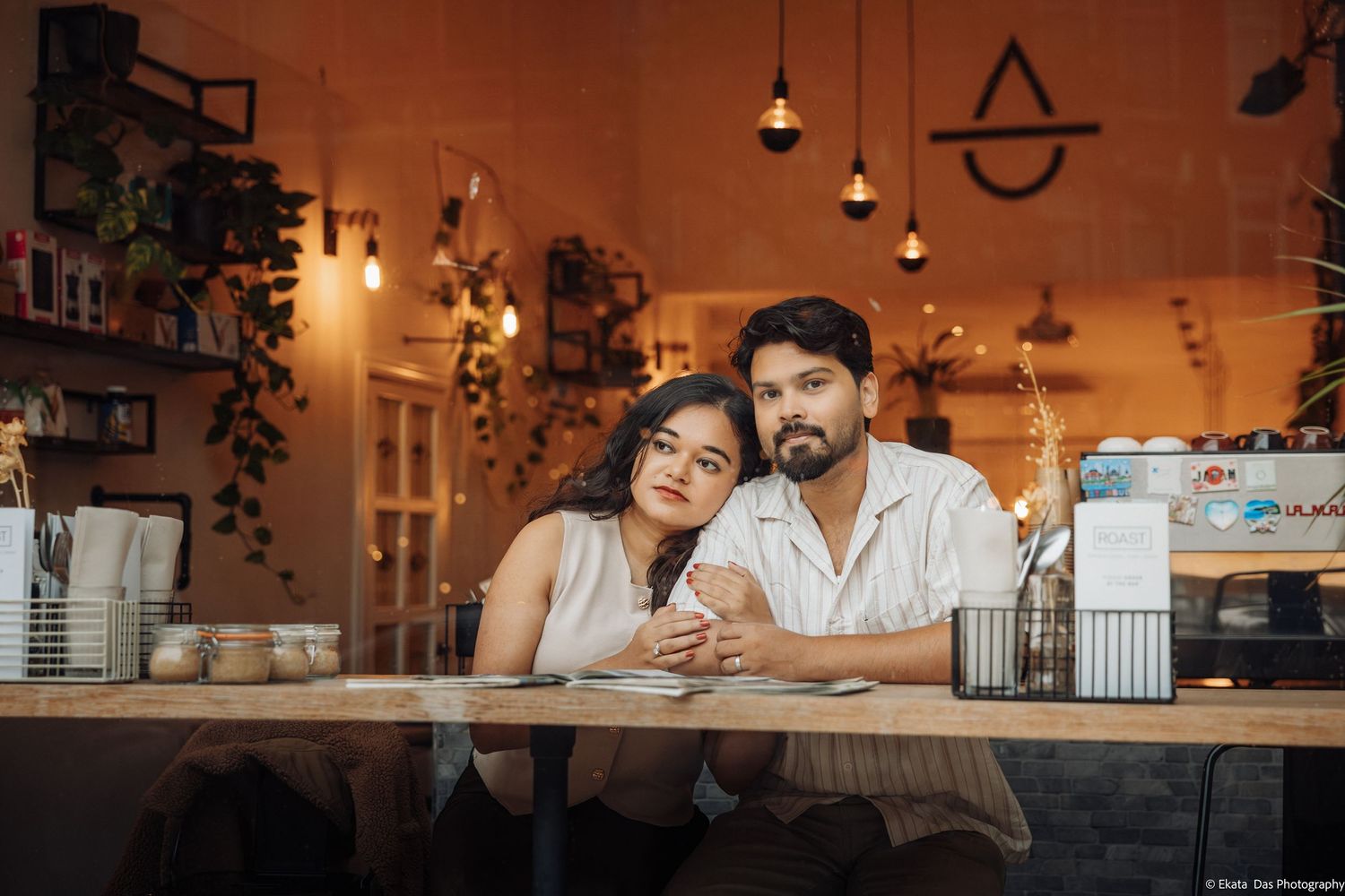 Smiling couple sitting close together at a wooden café table, surrounded by warm pendant lights, hanging plants and cozy shelves.
