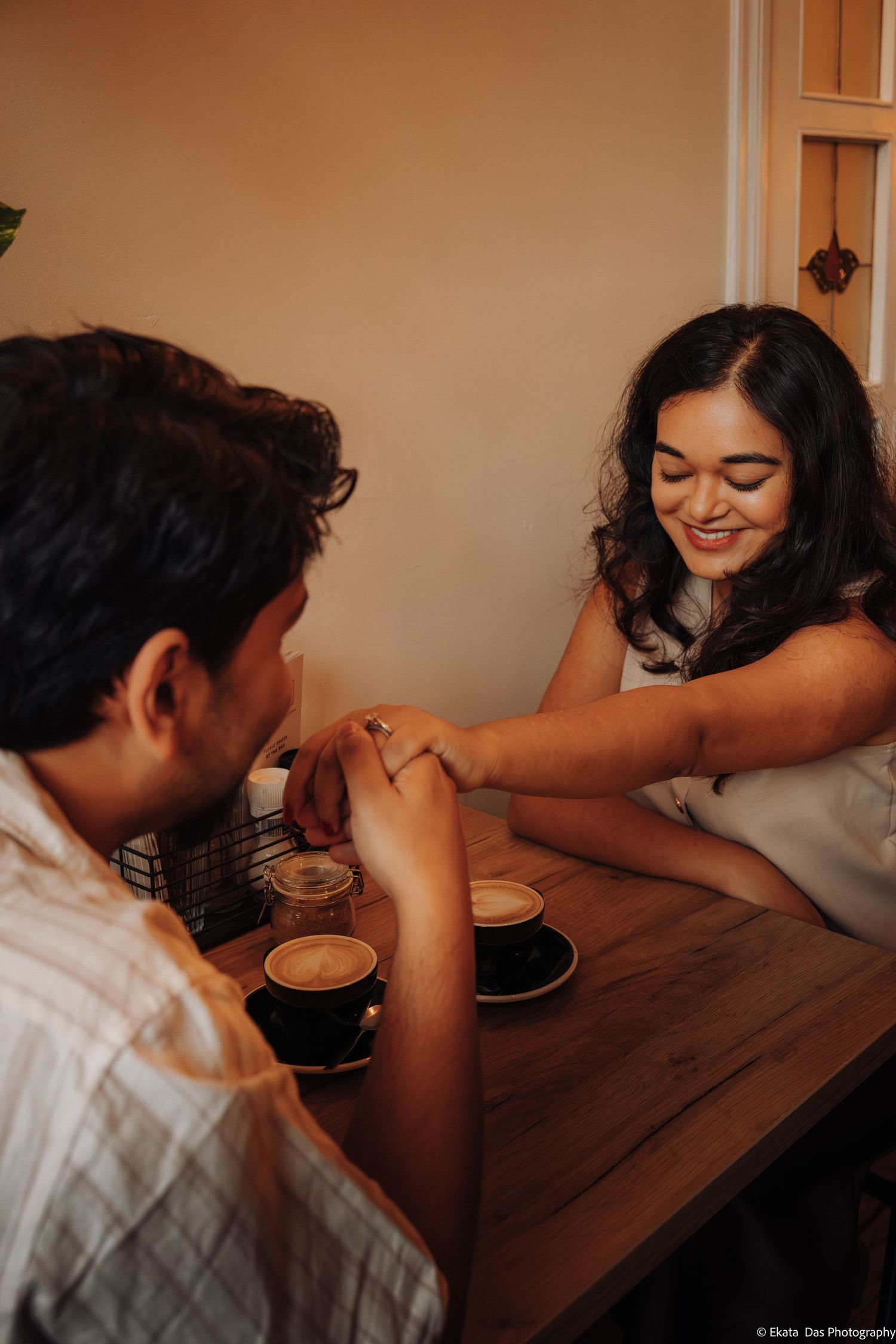 Smiling woman with long dark hair holds hands with a man across a wooden cafe table with two cups of latte art coffee.