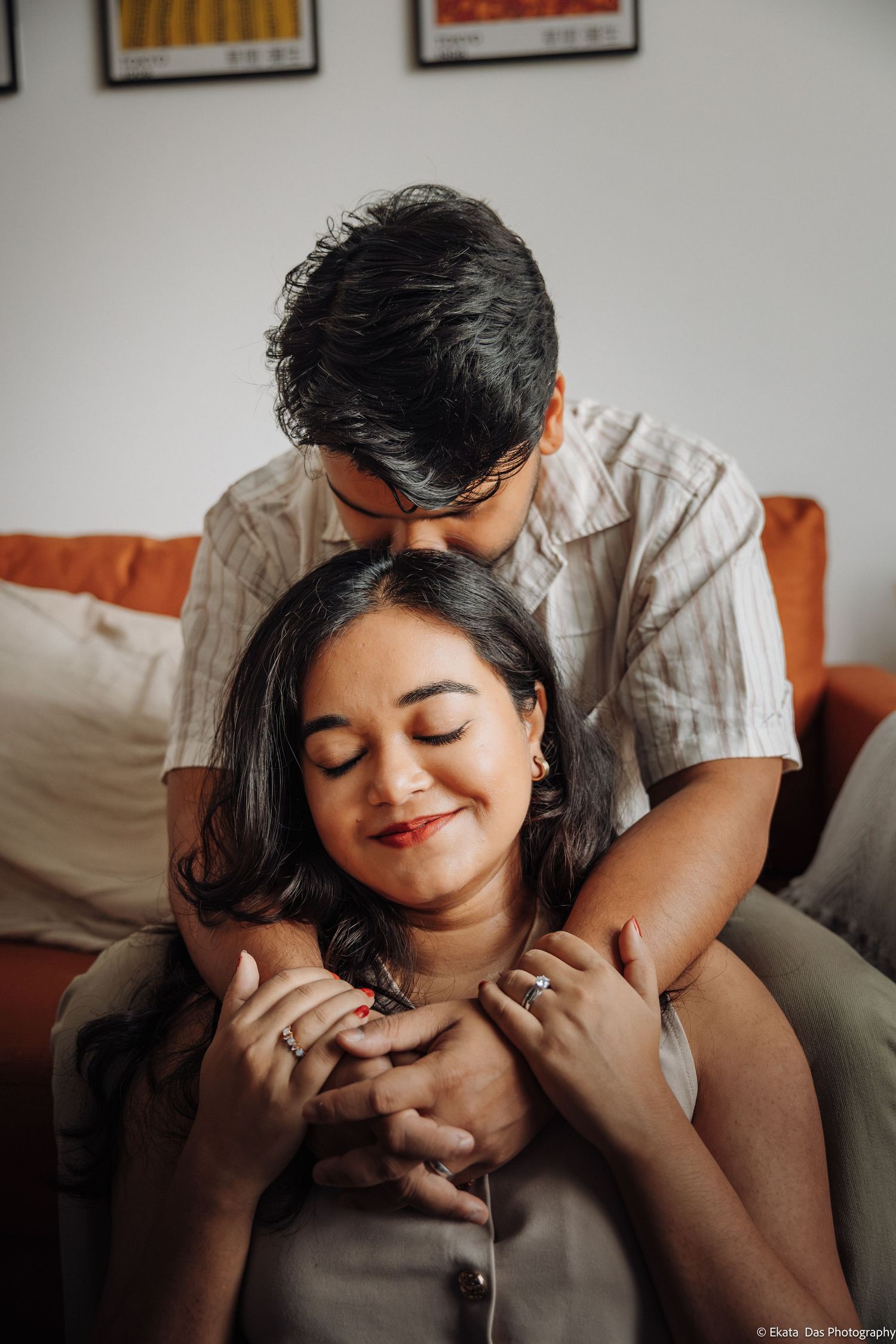 Smiling woman with closed eyes holds a man's hands as he leans over her shoulder in a tender couple portrait on an orange sofa.