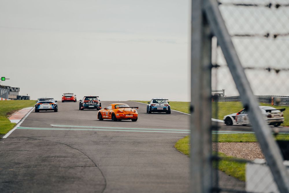 Race cars navigate a turn on a race track with an orange vehicle standing out among the pack of competitors.