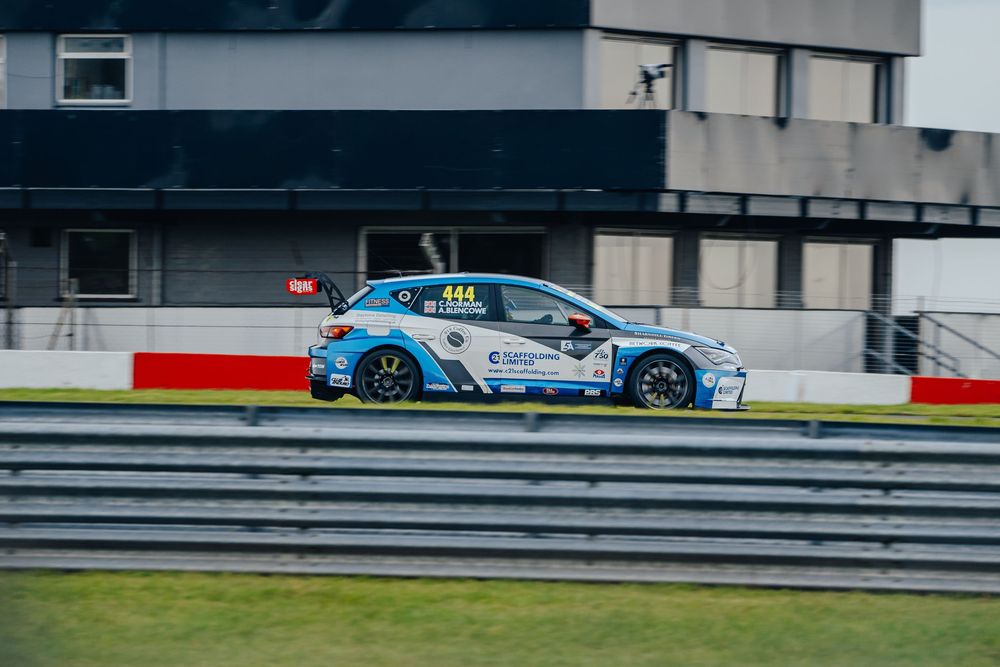 Blue racing car speeds along racetrack with white and red barriers during competitive motorsport event.