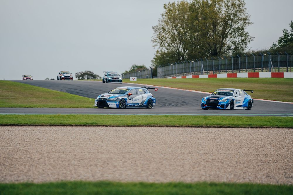 Racing cars compete on a professional circuit track with blue and white vehicles in action.