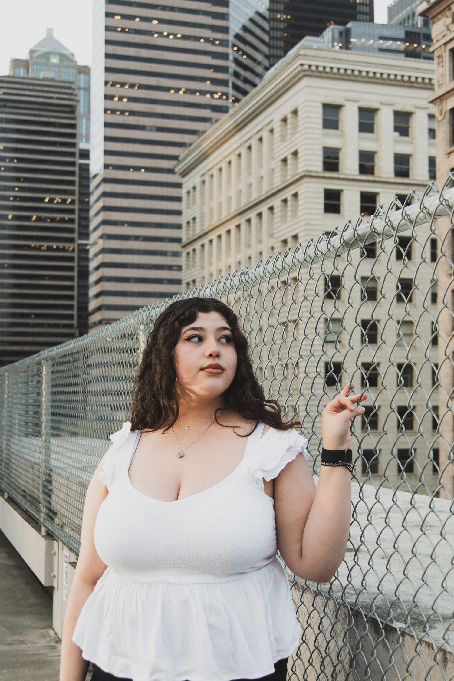 Woman in white dress poses against urban backdrop with modern buildings and chain link fence in series of photos.