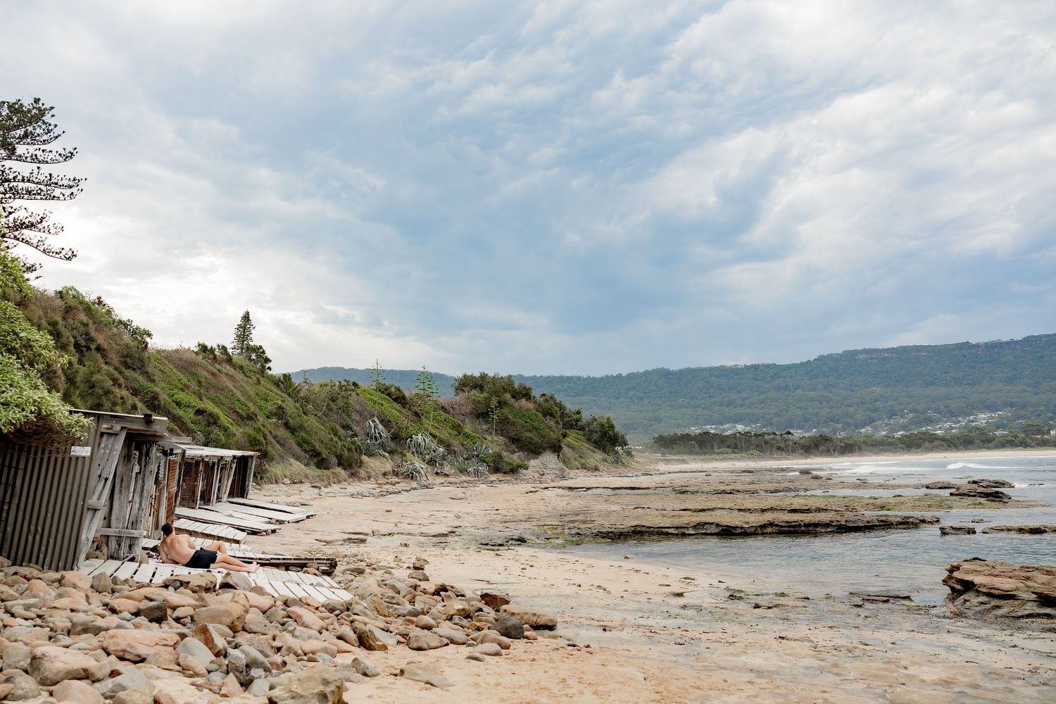 Panoramic coastal landscape showing rocky shoreline and calm waters with mountains in the distance under blue skies.