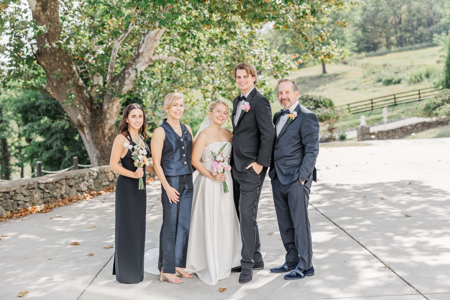 Wedding group photo with five people in formal attire standing outdoors under a tree.