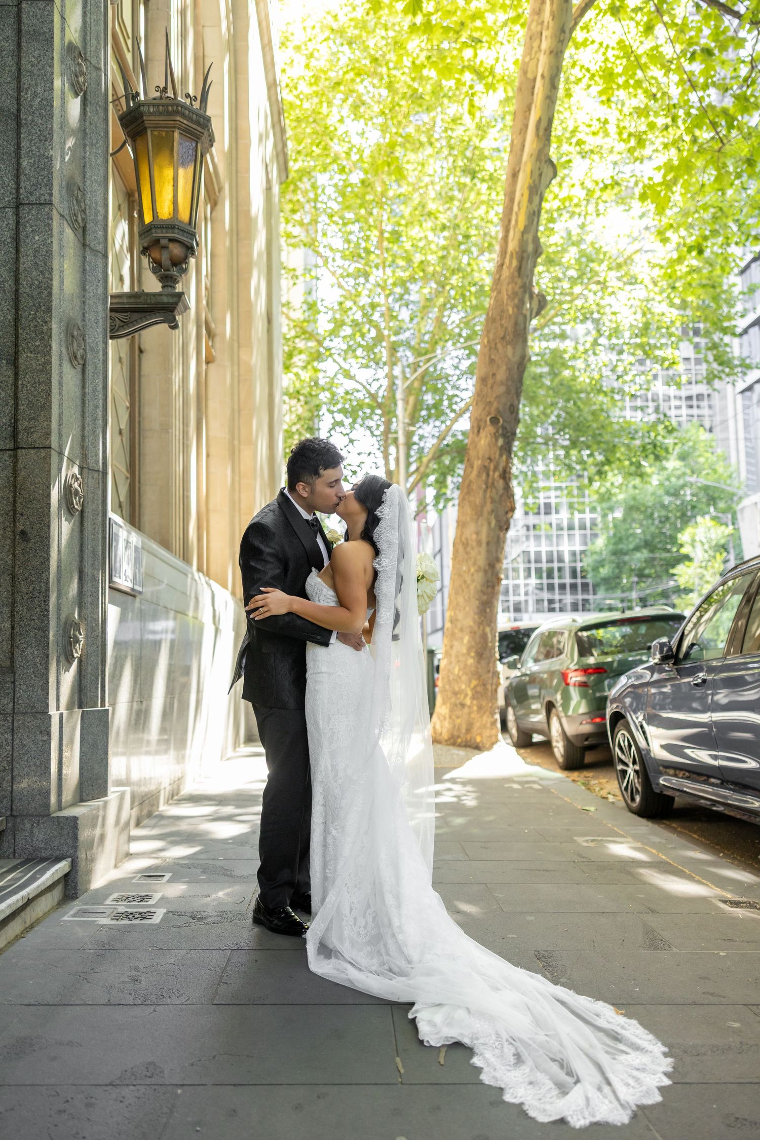 A couple in formal wedding attire embraces under a tree-lined urban street with parked cars in the background.