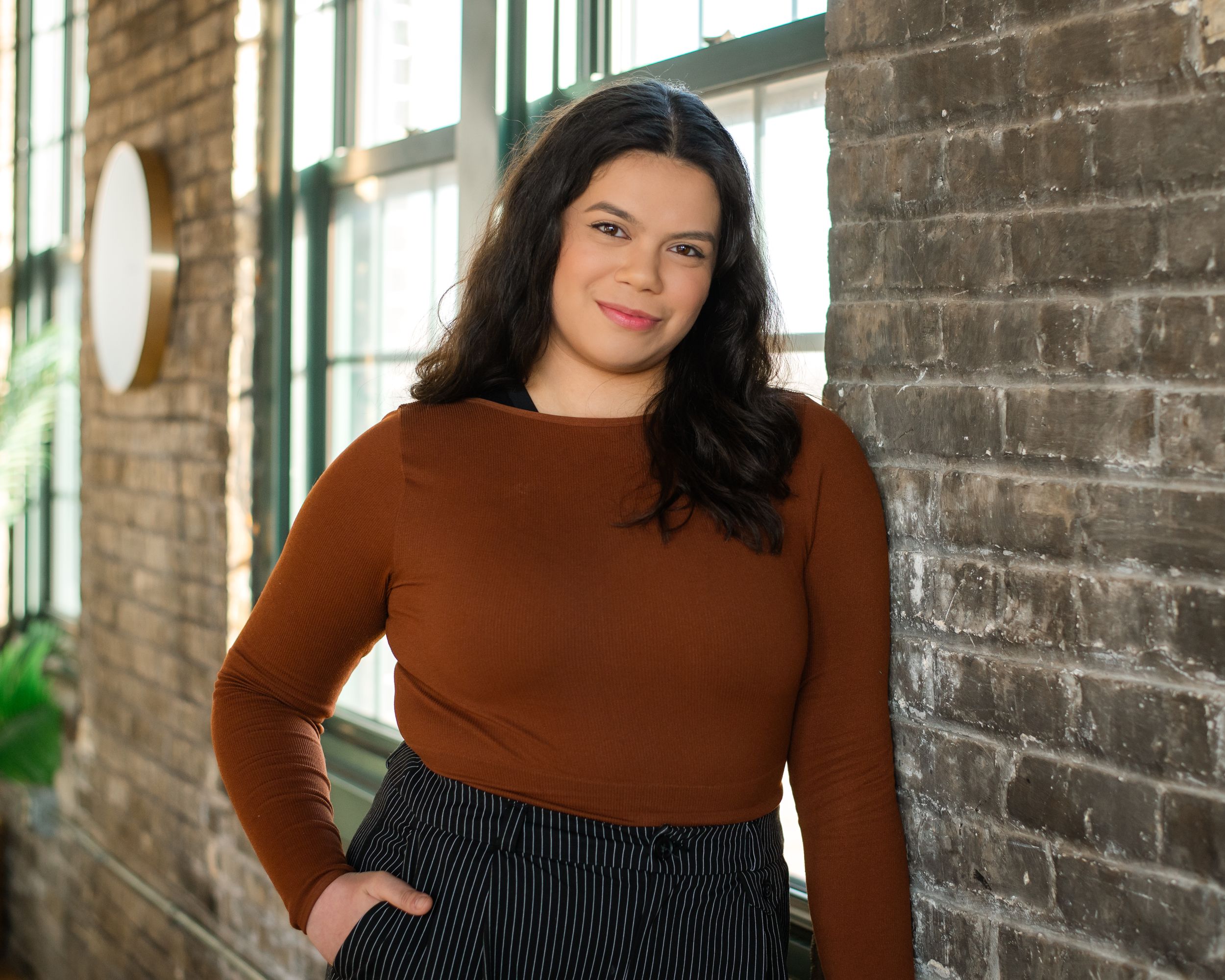 Someone poses in a brown sweater and striped skirt against a brick wall with natural lighting.