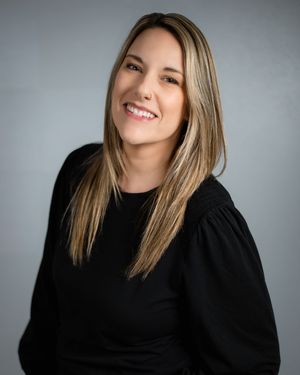 Professional portrait of a person with long blonde hair wearing a black top against a gray background, smiling warmly.