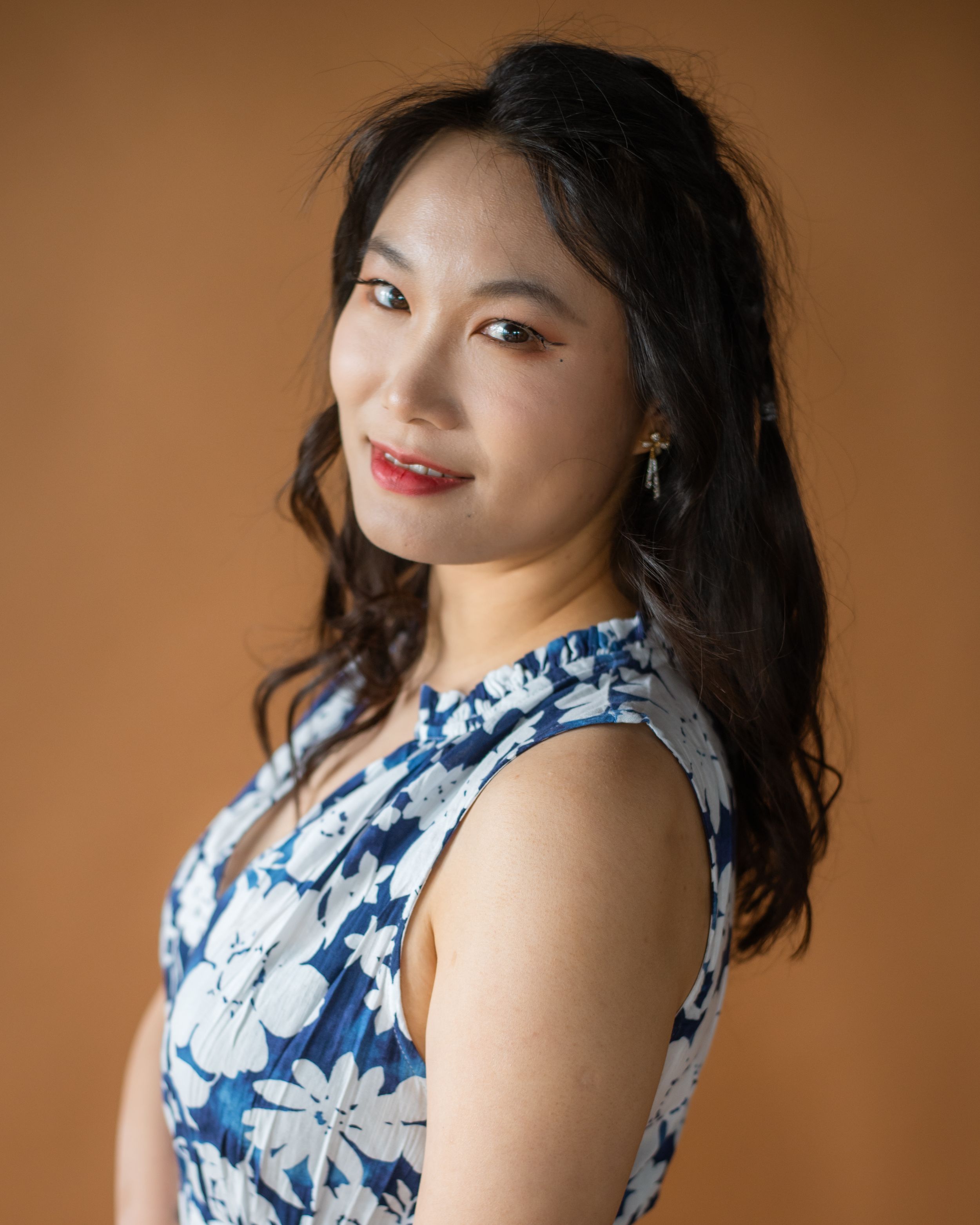 Woman in blue and white floral sleeveless dress poses against warm brown backdrop.