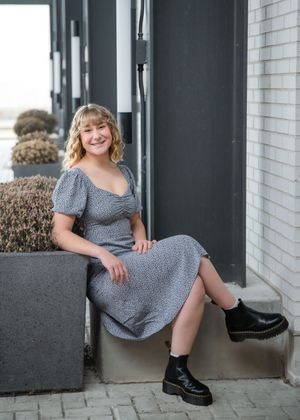 A person in a gray dress and black boots poses casually on a concrete ledge outside a brick building.