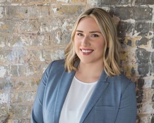 Professional in a blue blazer and white top poses against a rustic brick wall background.