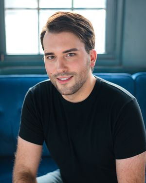 Person in black t-shirt sitting against blue background smiling warmly in casual portrait series.