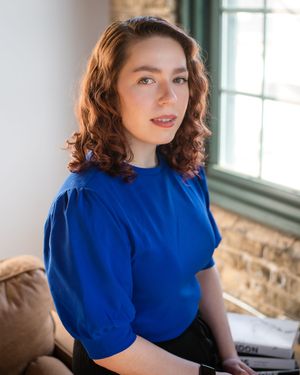 Professional portrait series in a bright room wearing a royal blue top against a window and stone wall backdrop.