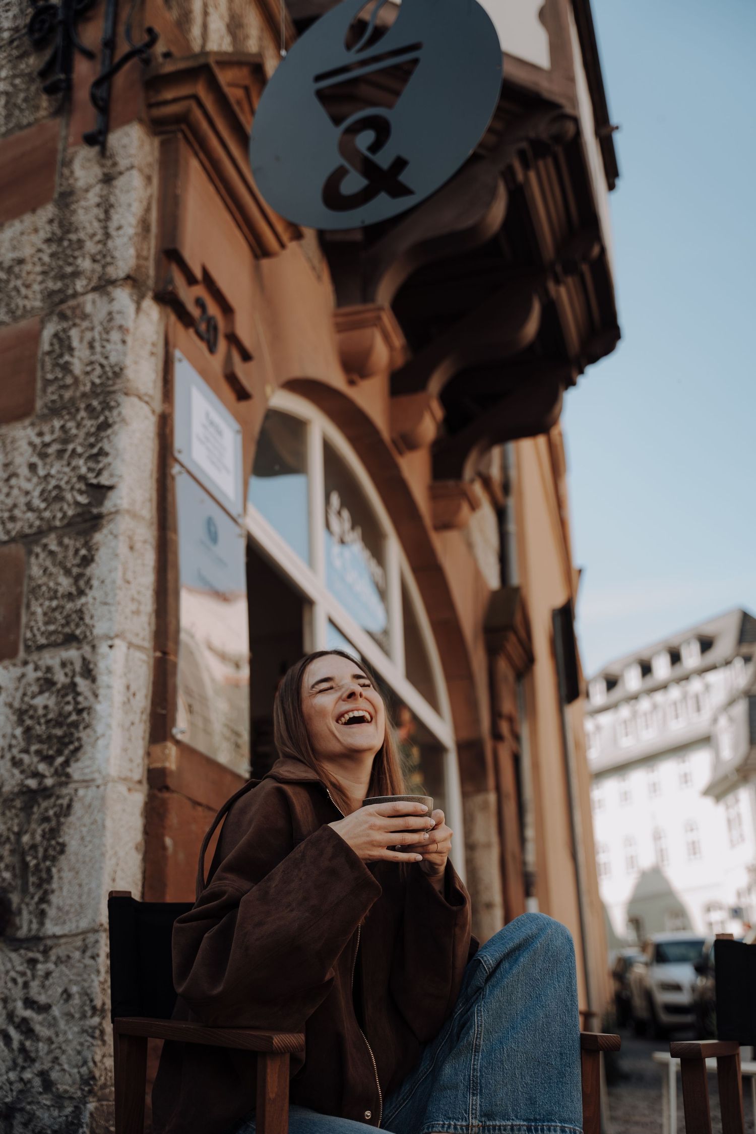 Fotografin / Frau sitzt vor einem malerischen Gebäude auf der Terrasse eines Cafés, trinkt einen Kaffee und lacht herzhaft dabei. Fotografie