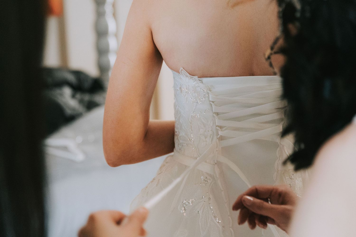 Close-up view of a white wedding dress being laced up in the back with delicate corset-style ribbons.