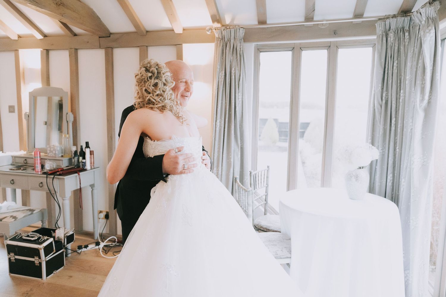 Wedding preparation scene showing someone adjusting a white bridal gown in a bright room with exposed wooden beams.