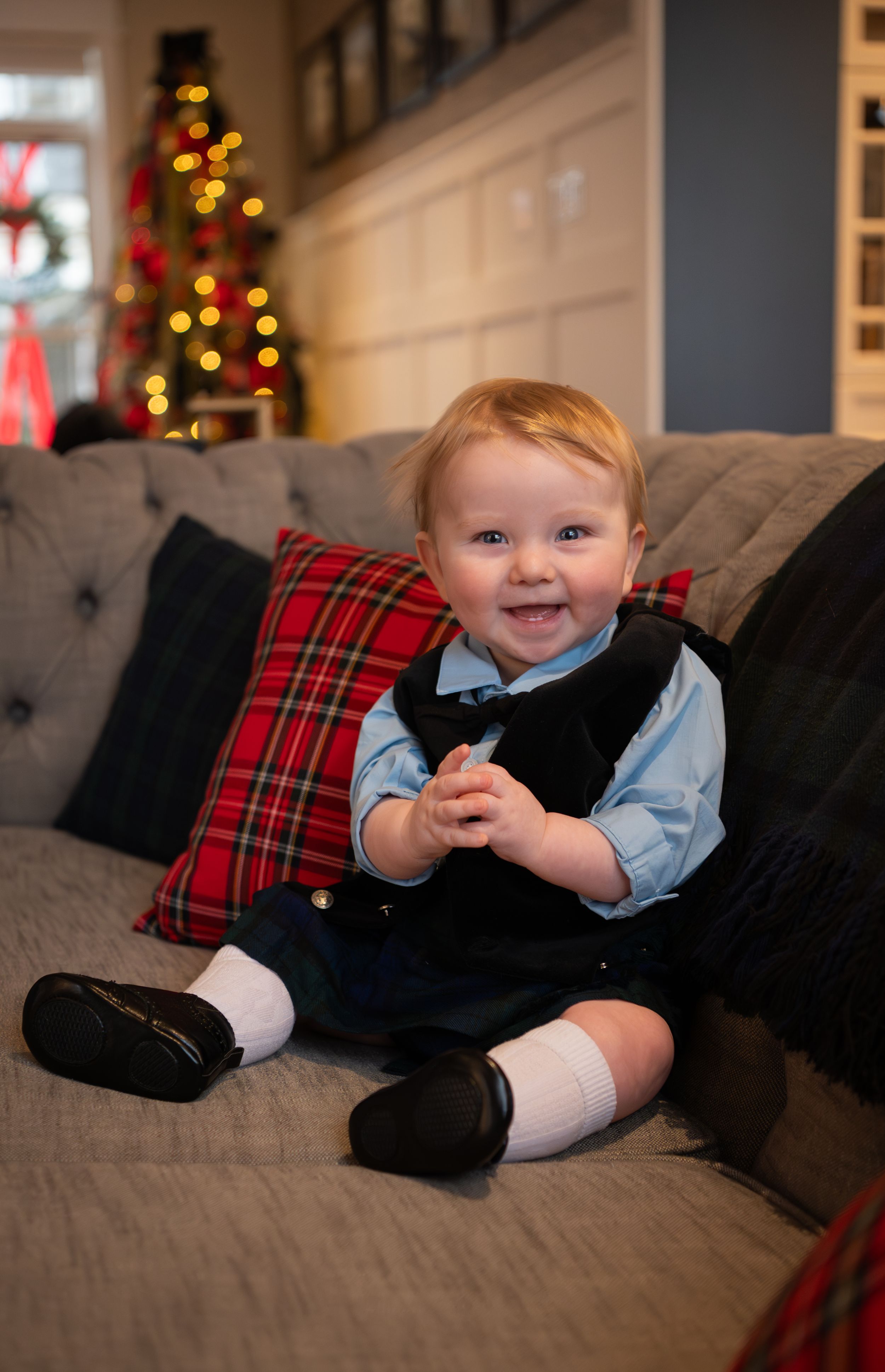 Baby in navy vest and blue shirt lying on gray couch with plaid pillows and Christmas lights in background.