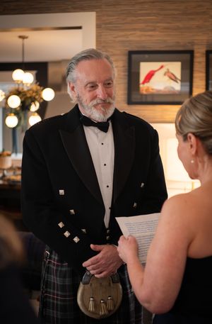 A person in a formal black tuxedo and Scottish kilt engages in conversation at an elegant indoor event.