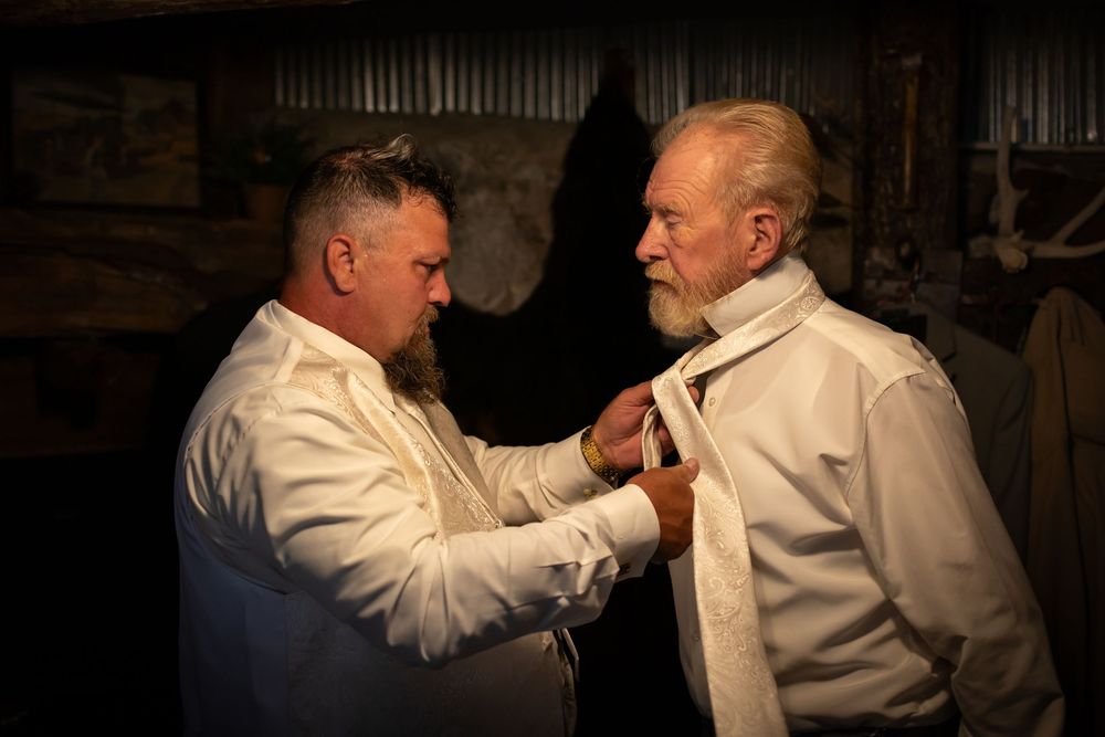 Two men in white dress shirts helping with formal attire preparations.