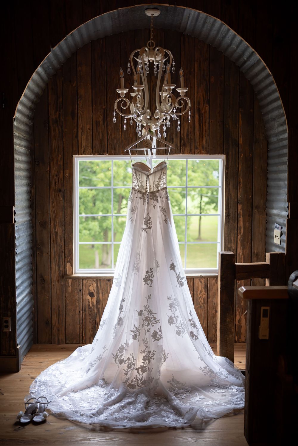 A white wedding dress with delicate lace details hangs elegantly in front of a rustic wooden window with natural light.