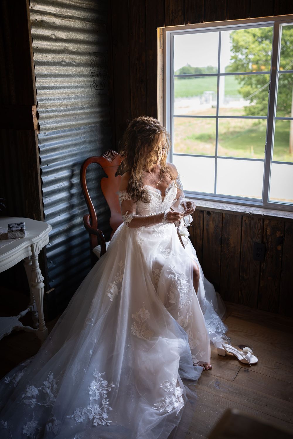 An elegant white wedding gown with flowing train is displayed in a rustic barn setting with natural window lighting.