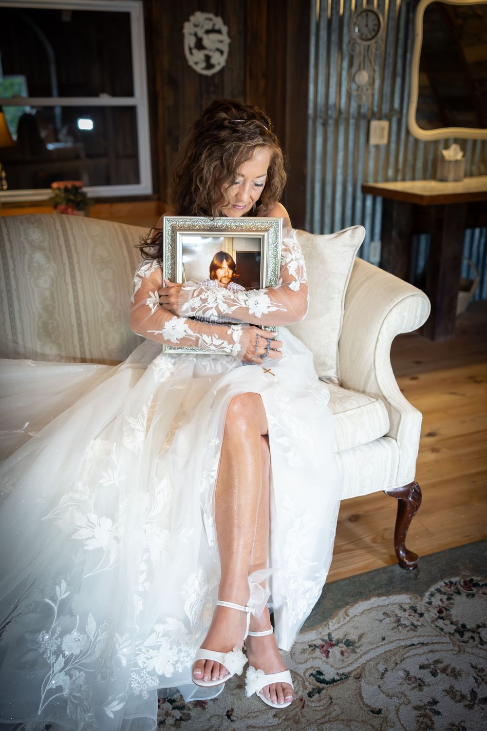 Bride in white dress sits on vintage chair holding a framed photograph.