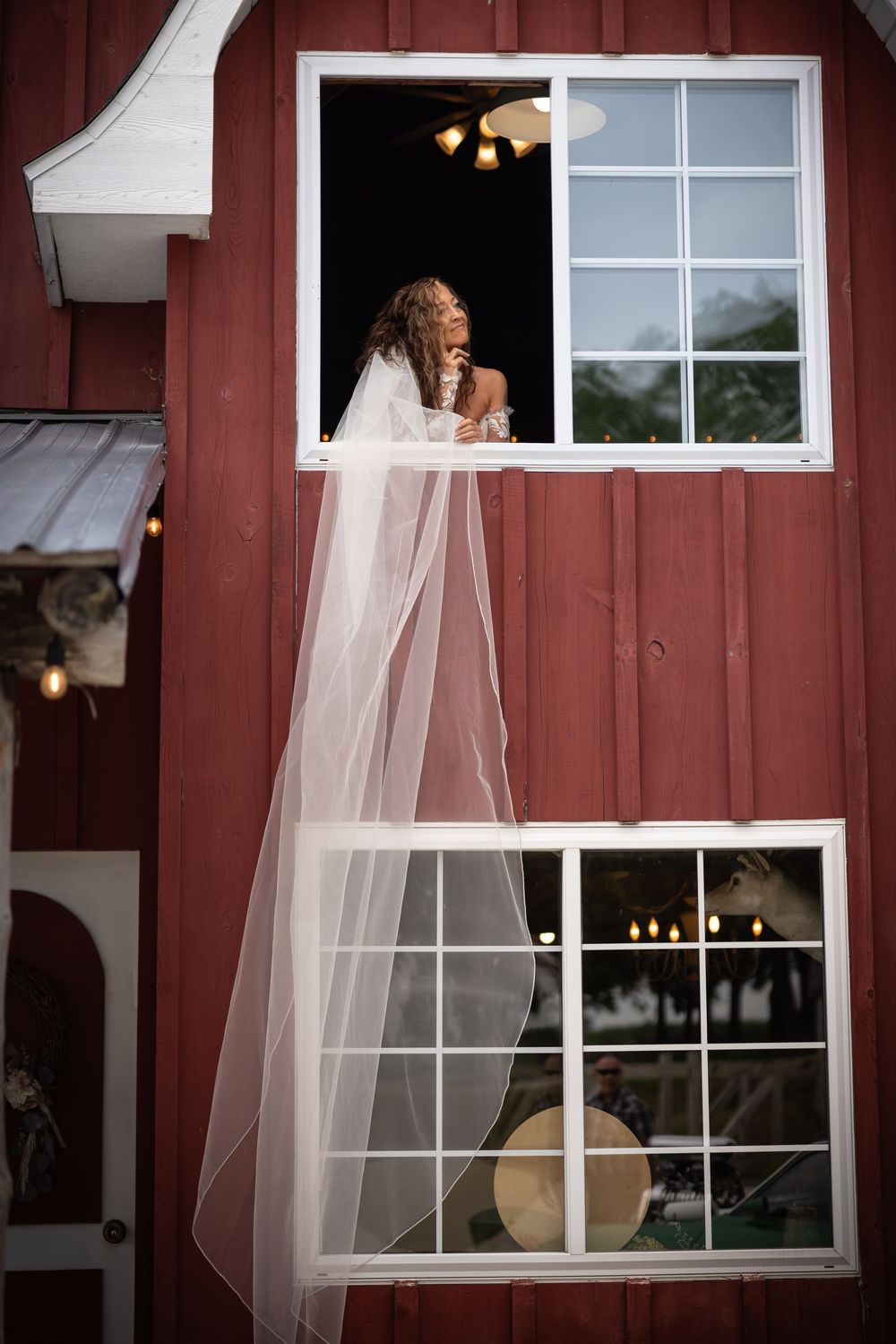 A flowing white wedding veil hangs from a rustic red barn window creating an ethereal display.