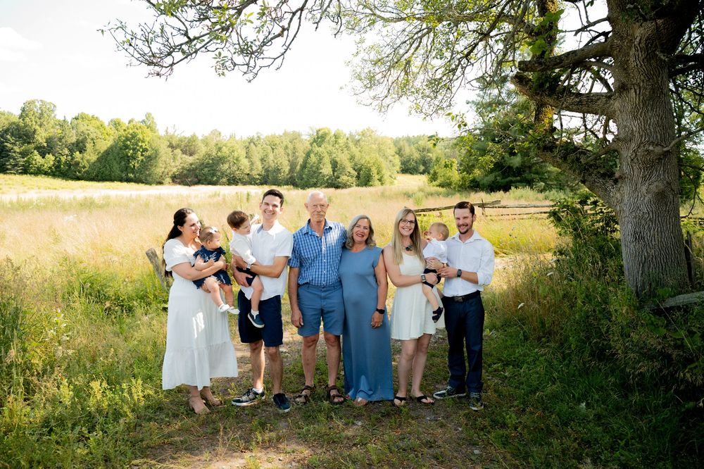 A group of people gather for a casual outdoor photo in a sunny meadow with trees and tall grass in the background.