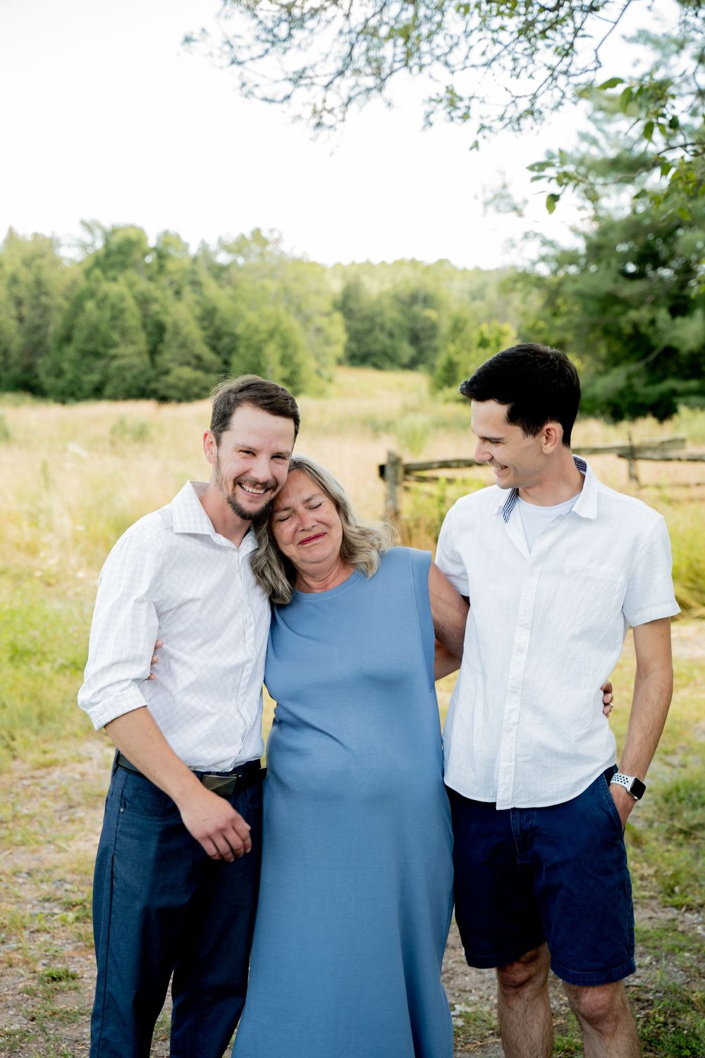 Three people share a candid moment together outdoors in a grassy field on a sunny day.