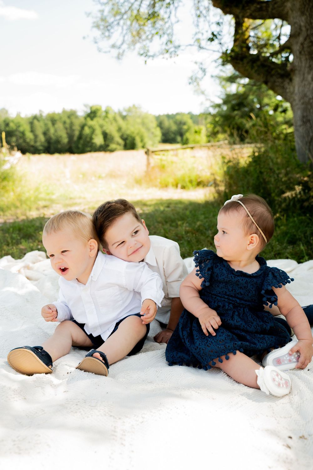 Babies in white and navy outfits lying on a white blanket under a tree.