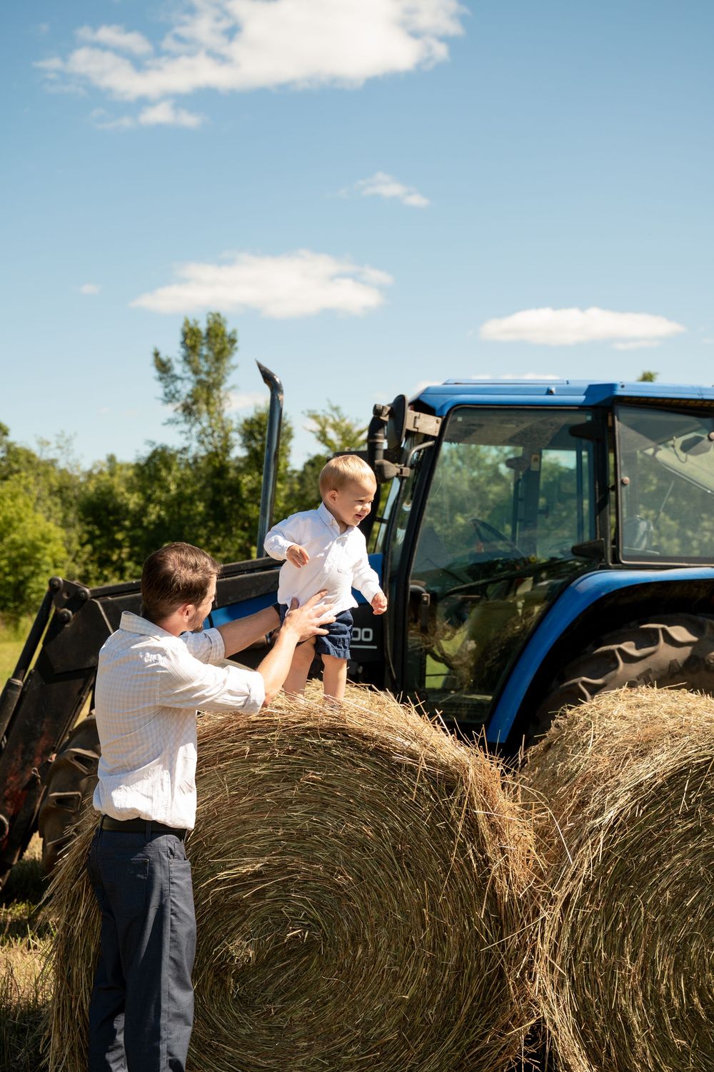Children playing and climbing on large round hay bales next to a blue tractor in a sunny field.
