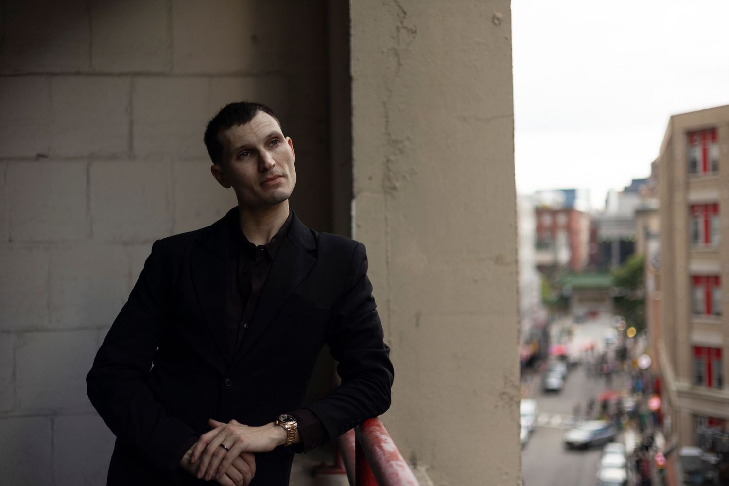 Person in a black coat stands on a balcony overlooking an urban street view in moody lighting.