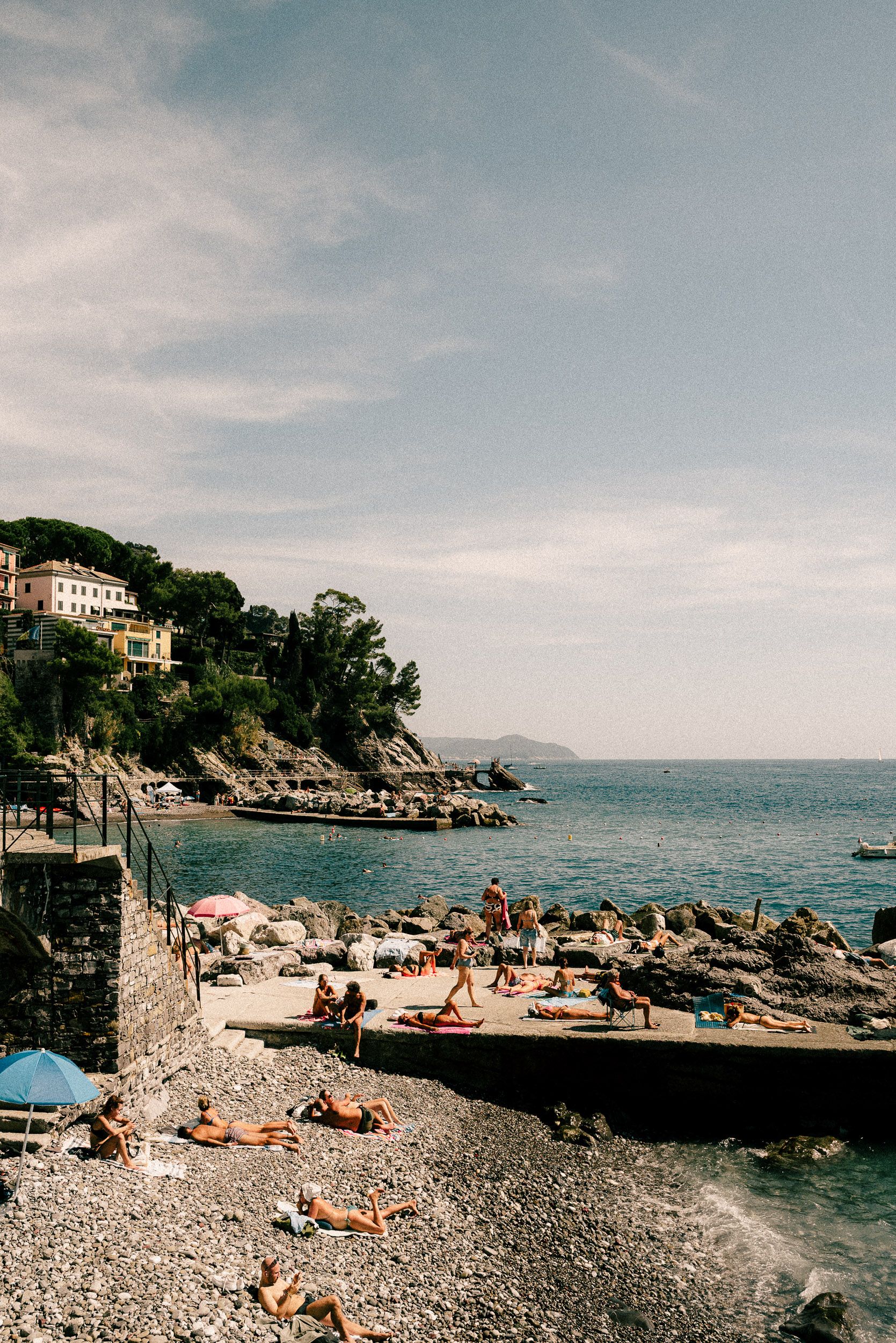 Sunbathers relax on a rocky Mediterranean beach cove with cliffside villas and boats in the distance.
