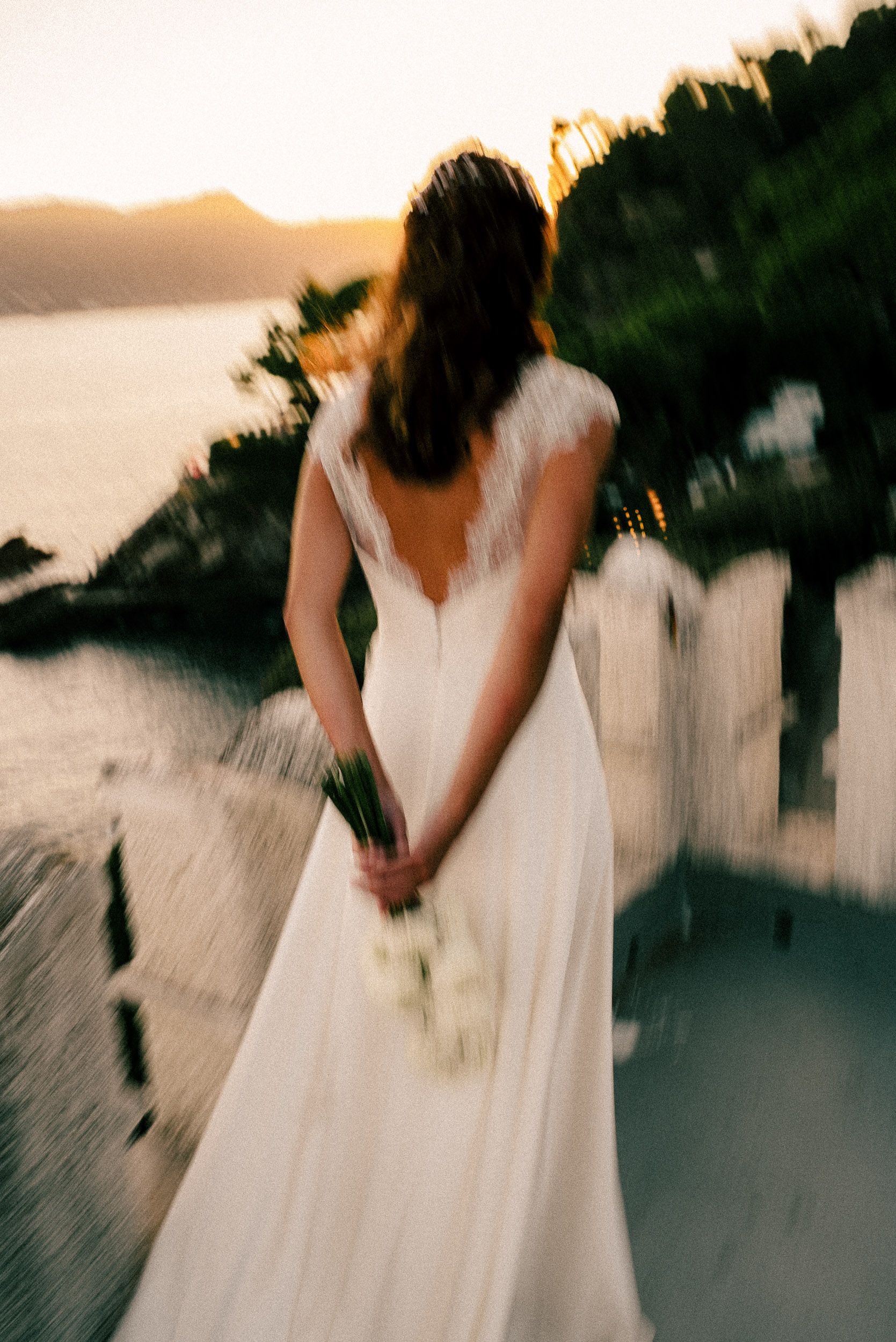 A bride in a flowing white backless dress stands at sunset overlooking a coastal vista.