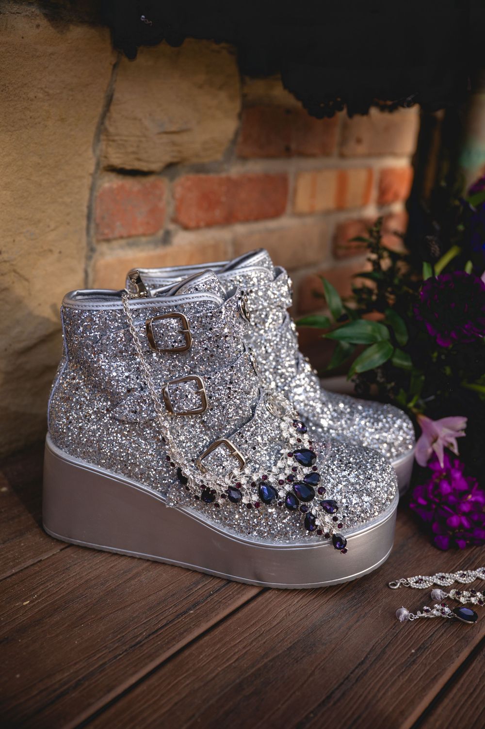 Sparkly silver platform ankle boot with buckle details sits on wooden surface next to purple flowers and brick wall.