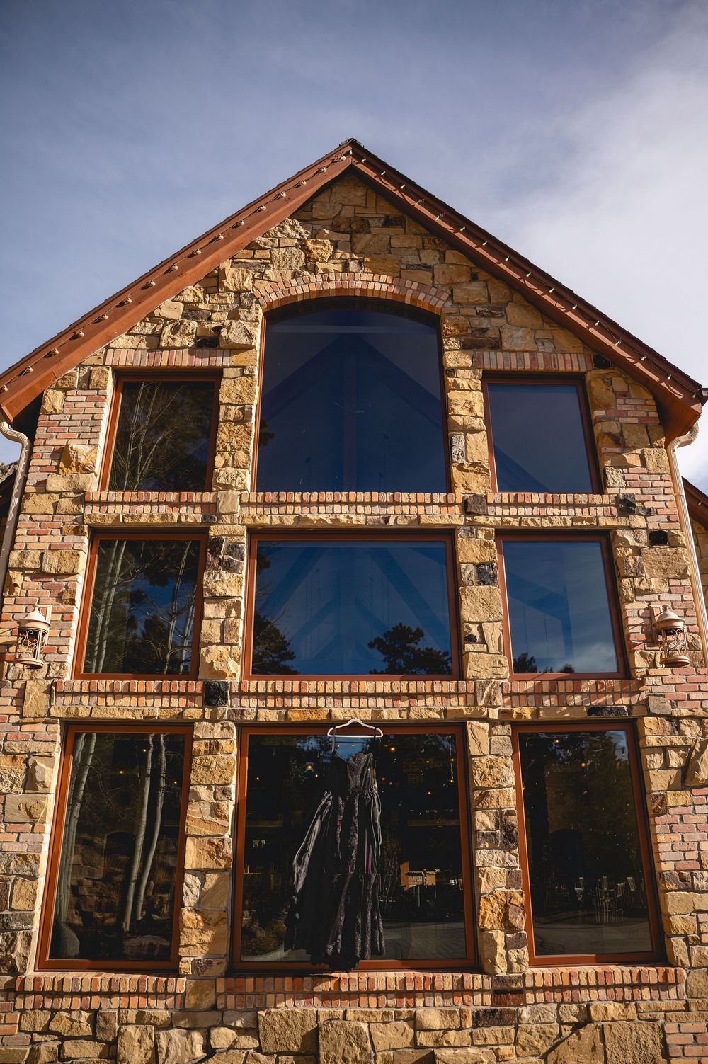 Stone facade of rustic house with three rows of large windows reflecting blue sky and trees in architectural design.