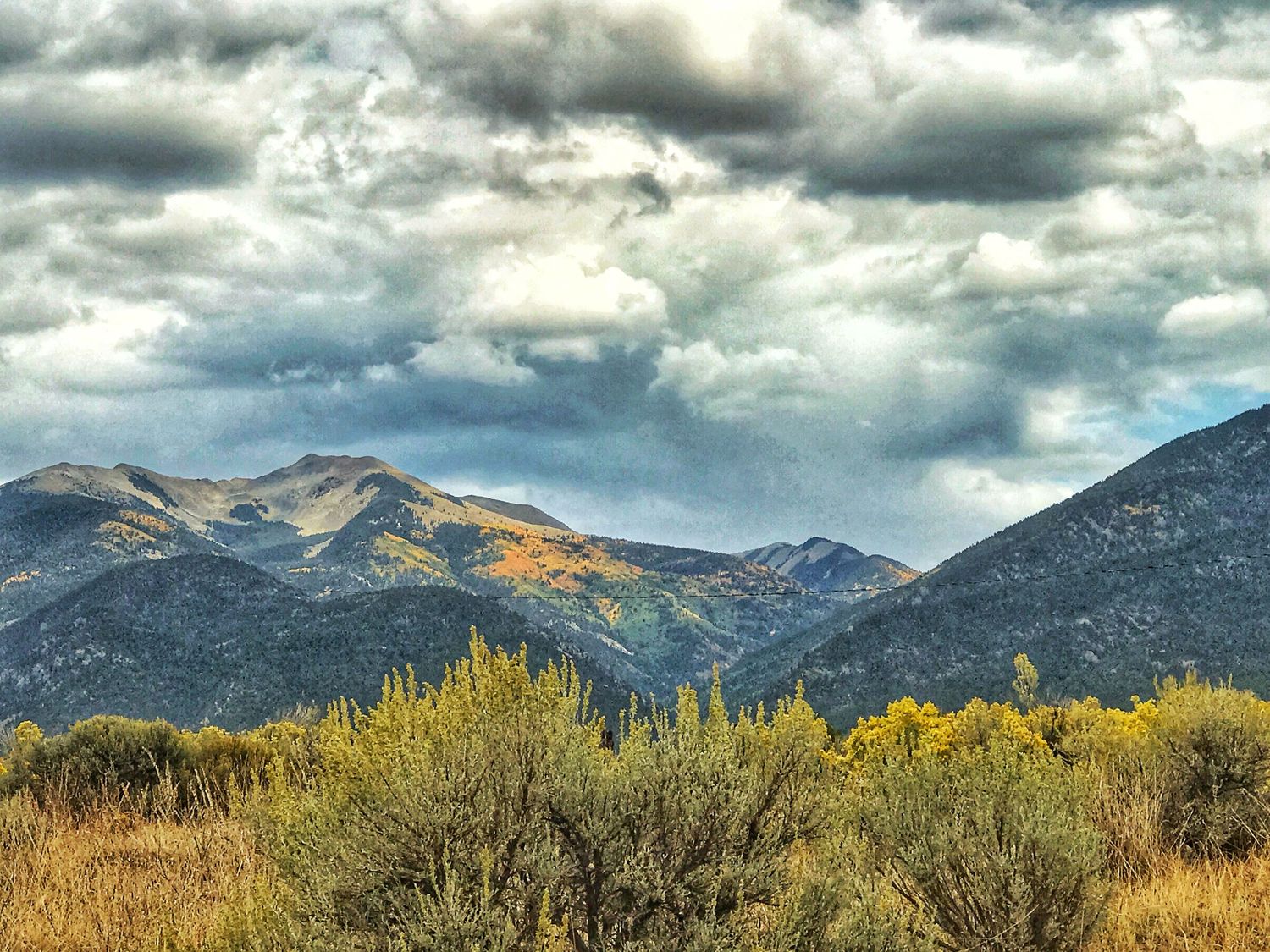 Dramatic cloudy skies over mountain peaks with sunlit autumn foliage in the foreground.