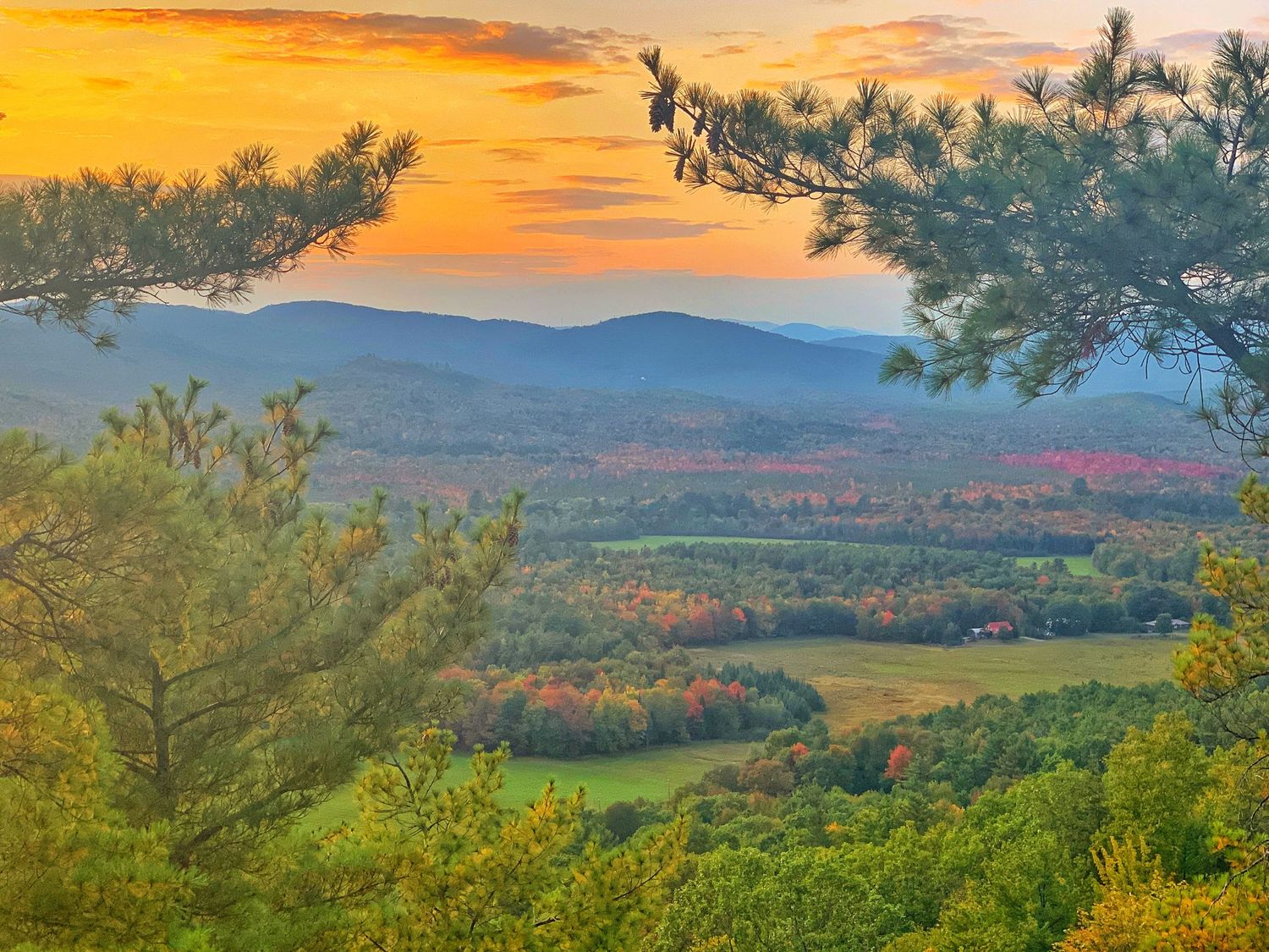 Dramatic orange sunset illuminates mountain valley with autumn foliage and distant peaks.