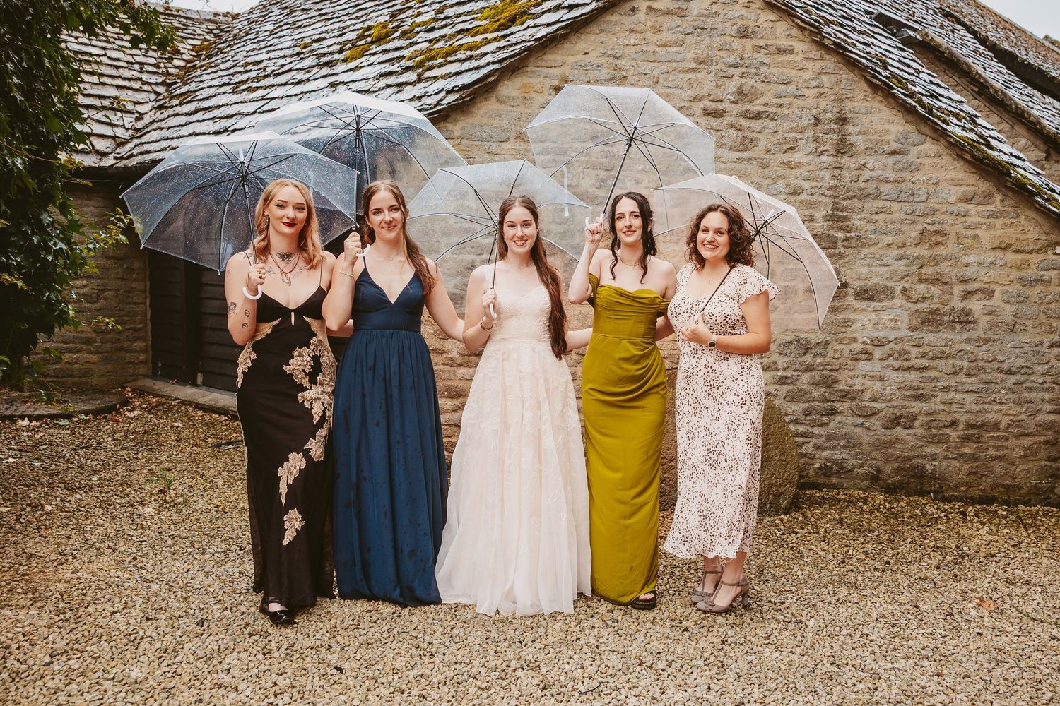 A group stands together against a rustic stone wall at a formal event wearing elegant dresses in various colors.