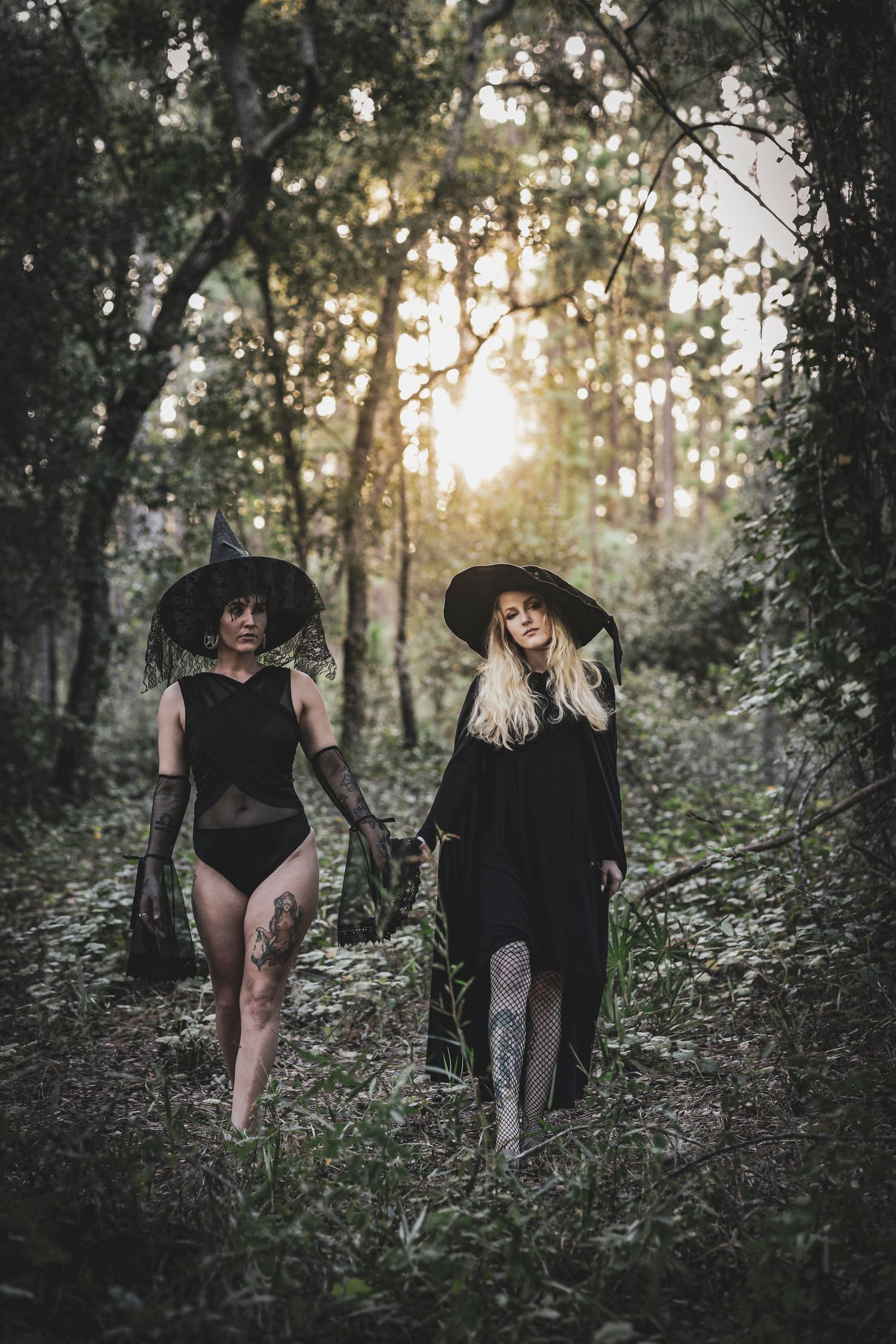 Two women in witch costumes walking through a forest at sunset.