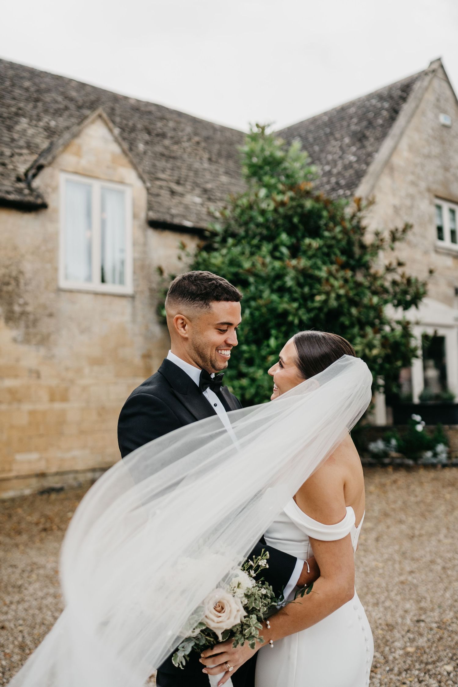 A romantic wedding couple embraces outdoors against a rustic stone building as the bride's veil flows in the wind.