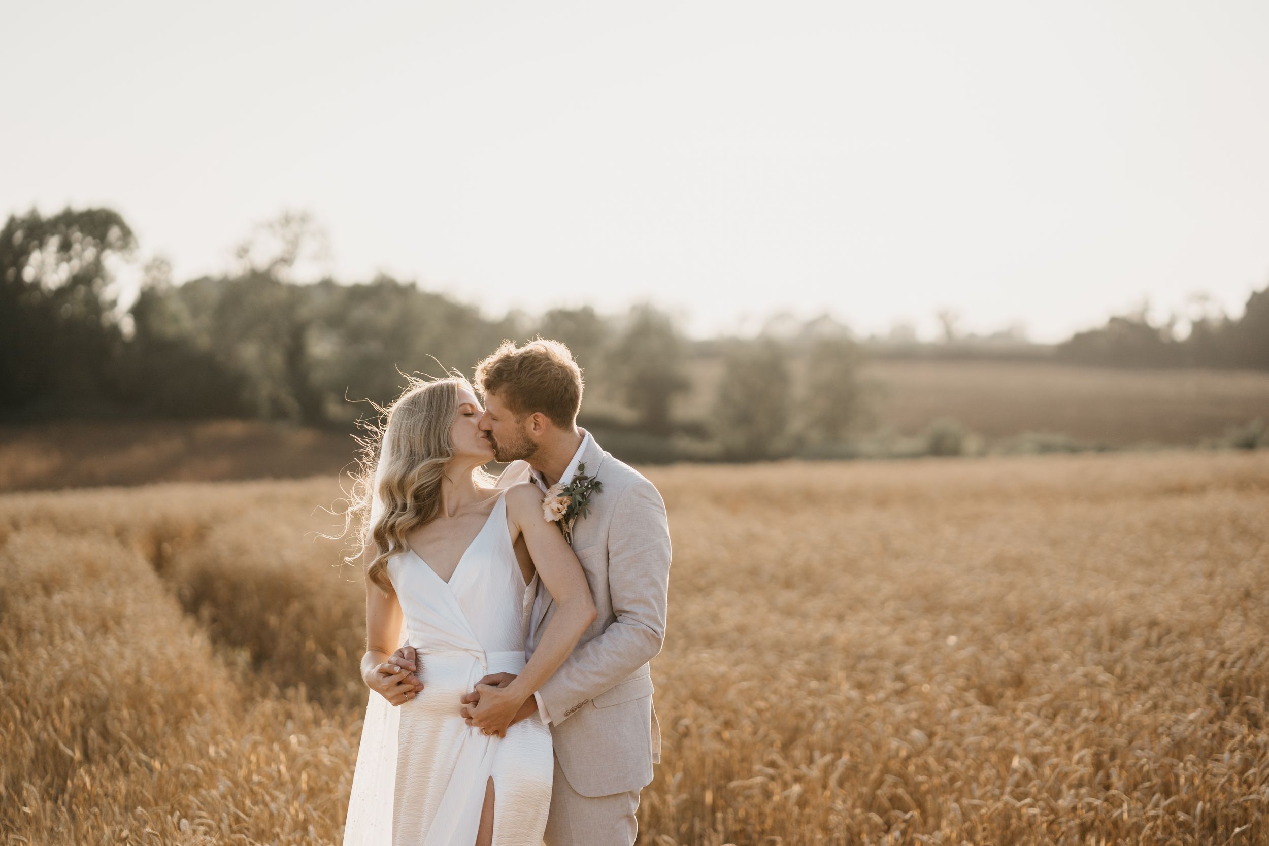 A romantic couple embraces in a golden wheat field at sunset during a rustic outdoor photoshoot.