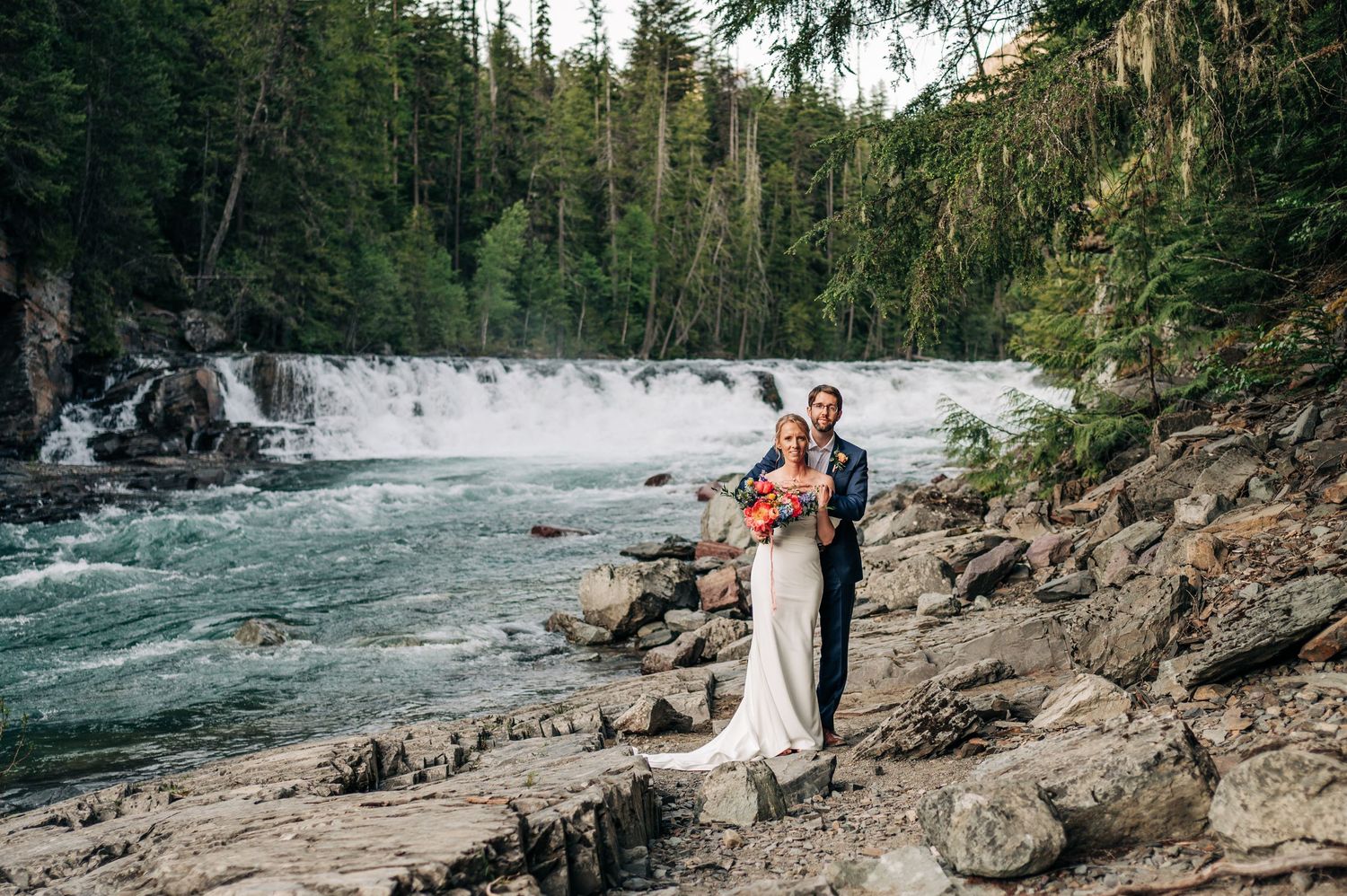 A couple embraces by a cascading waterfall in a forest setting with rocky shores and rushing turquoise waters.