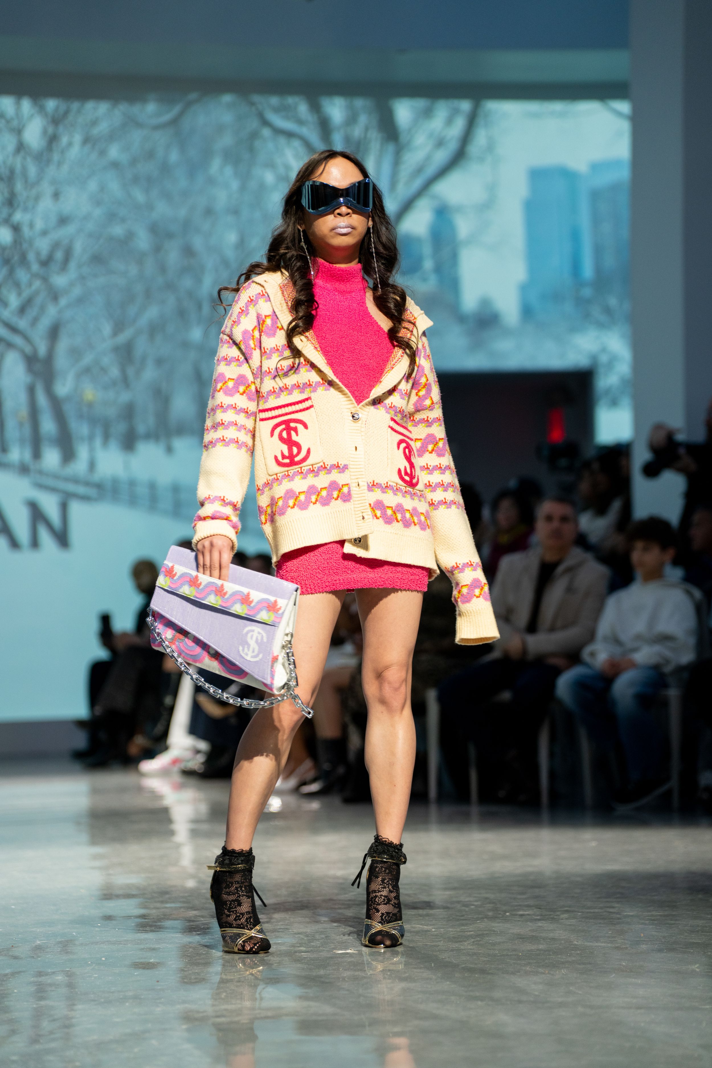 Model walks runway in pink mini skirt, patterned cardigan, sunglasses, and lace boots carrying colorful handbag at fashion show.