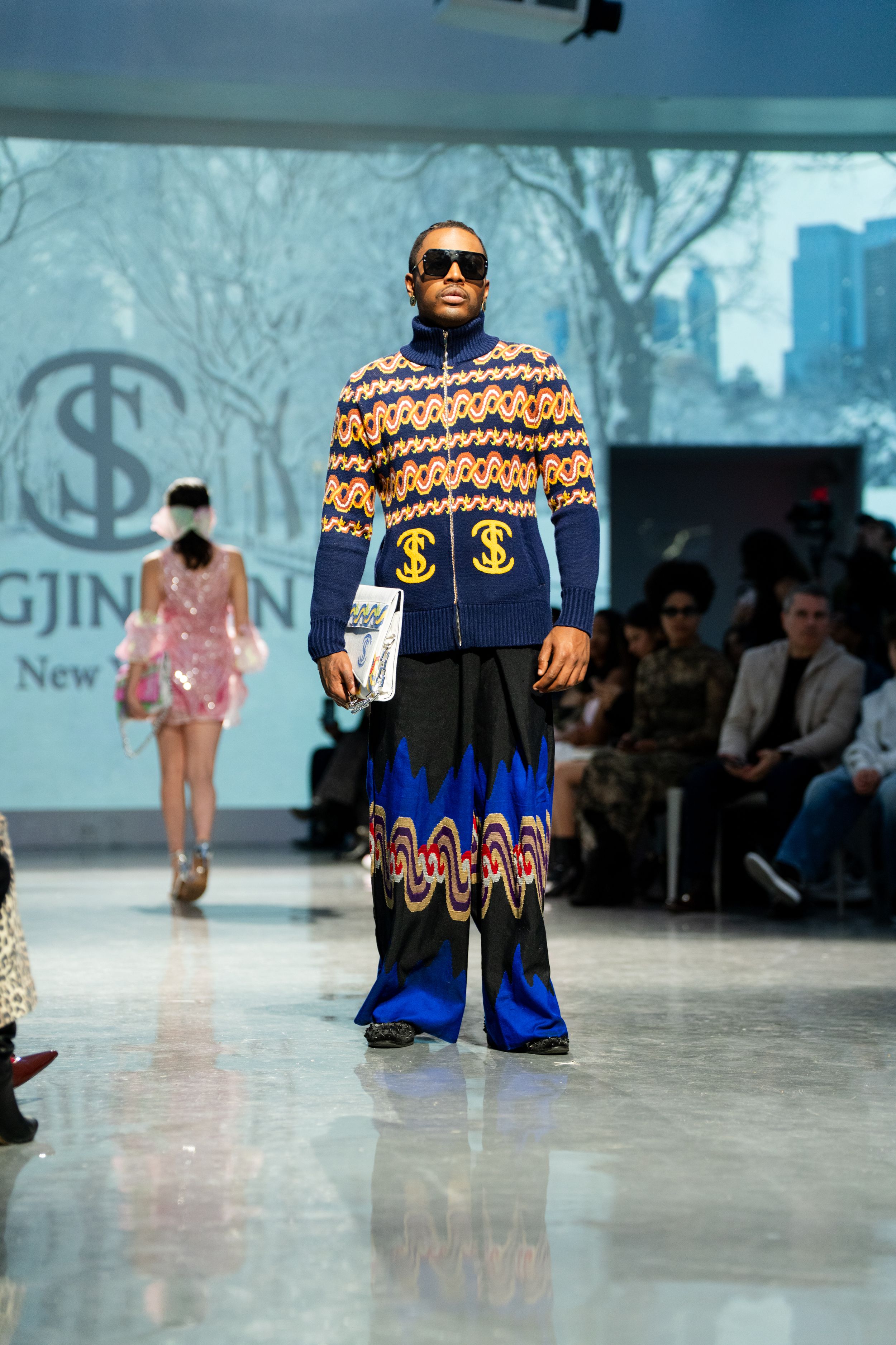 Model walks runway in bold patterned blue jacket with dollar signs and vibrant flame-print wide-leg pants at fashion show.