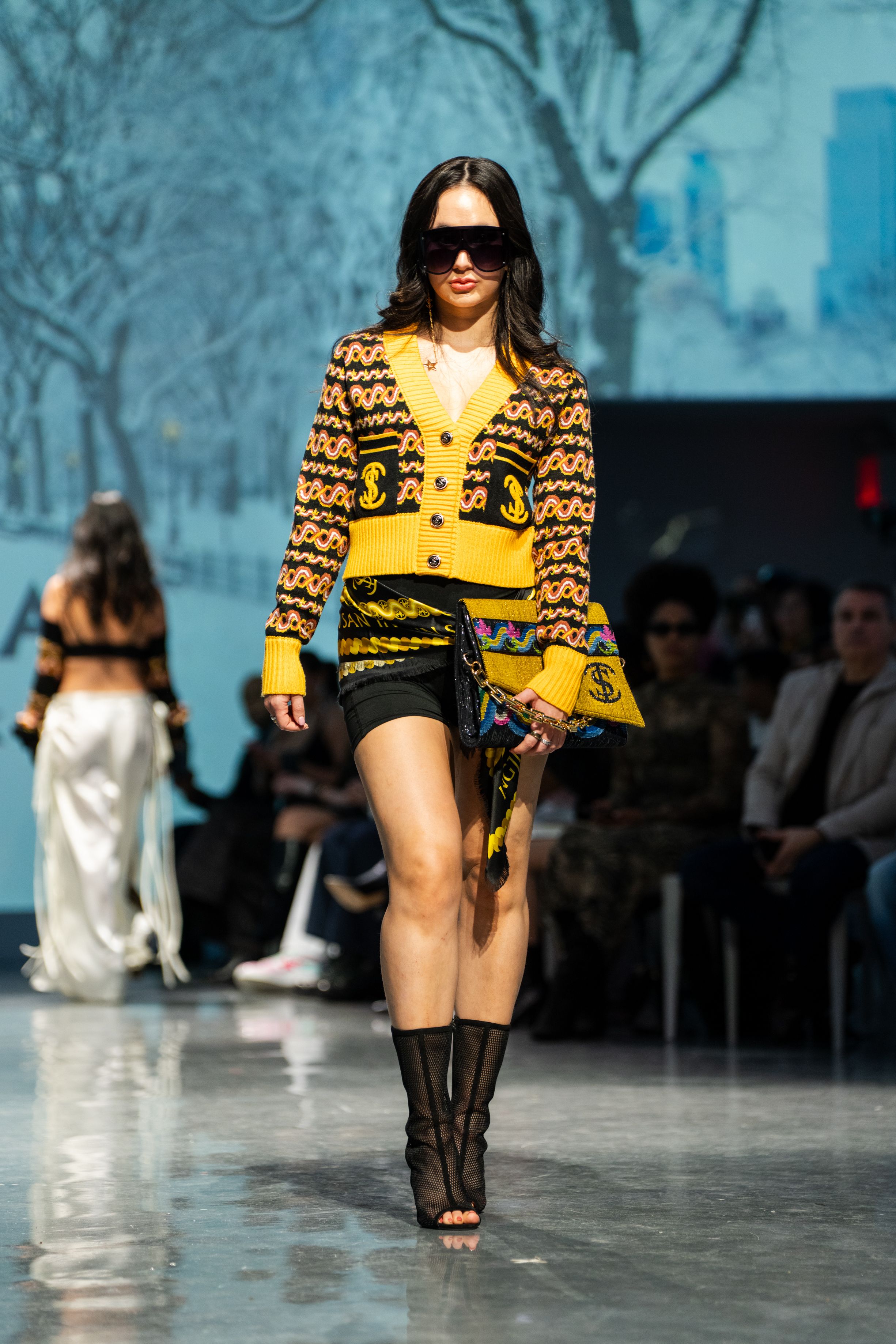 Model walks runway in yellow patterned cardigan, black shorts, and knee-high boots at a fashion show.