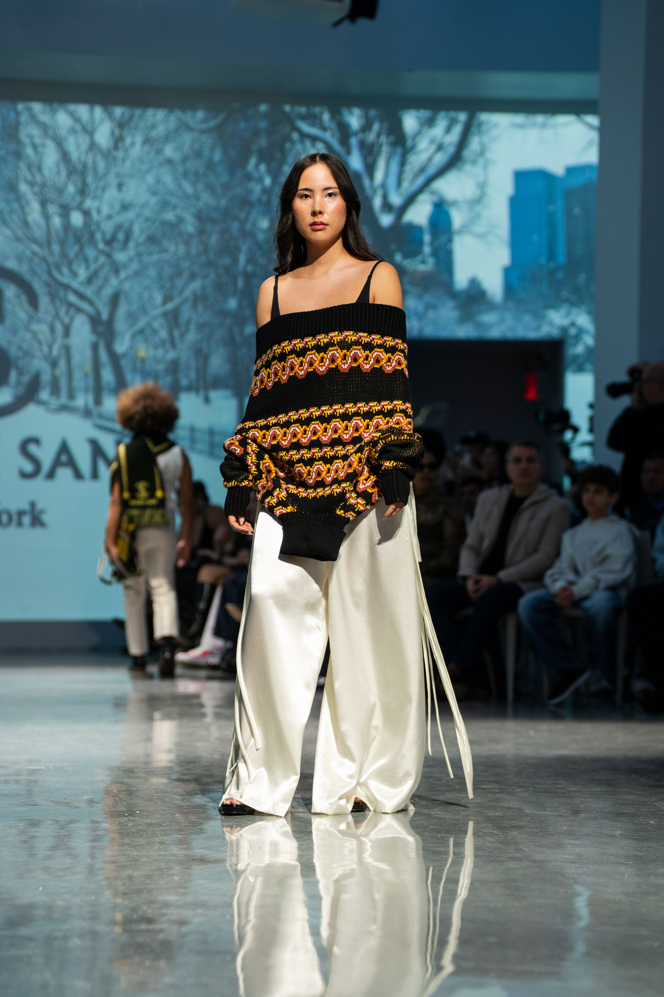 Model walks runway in black crochet sweater with orange accents and wide-leg white pants at a fashion show.