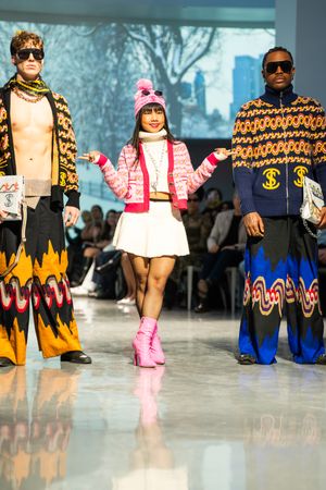 Three models walk a fashion runway wearing bold, colorful outfits with graphic prints and accessories at a fashion show.