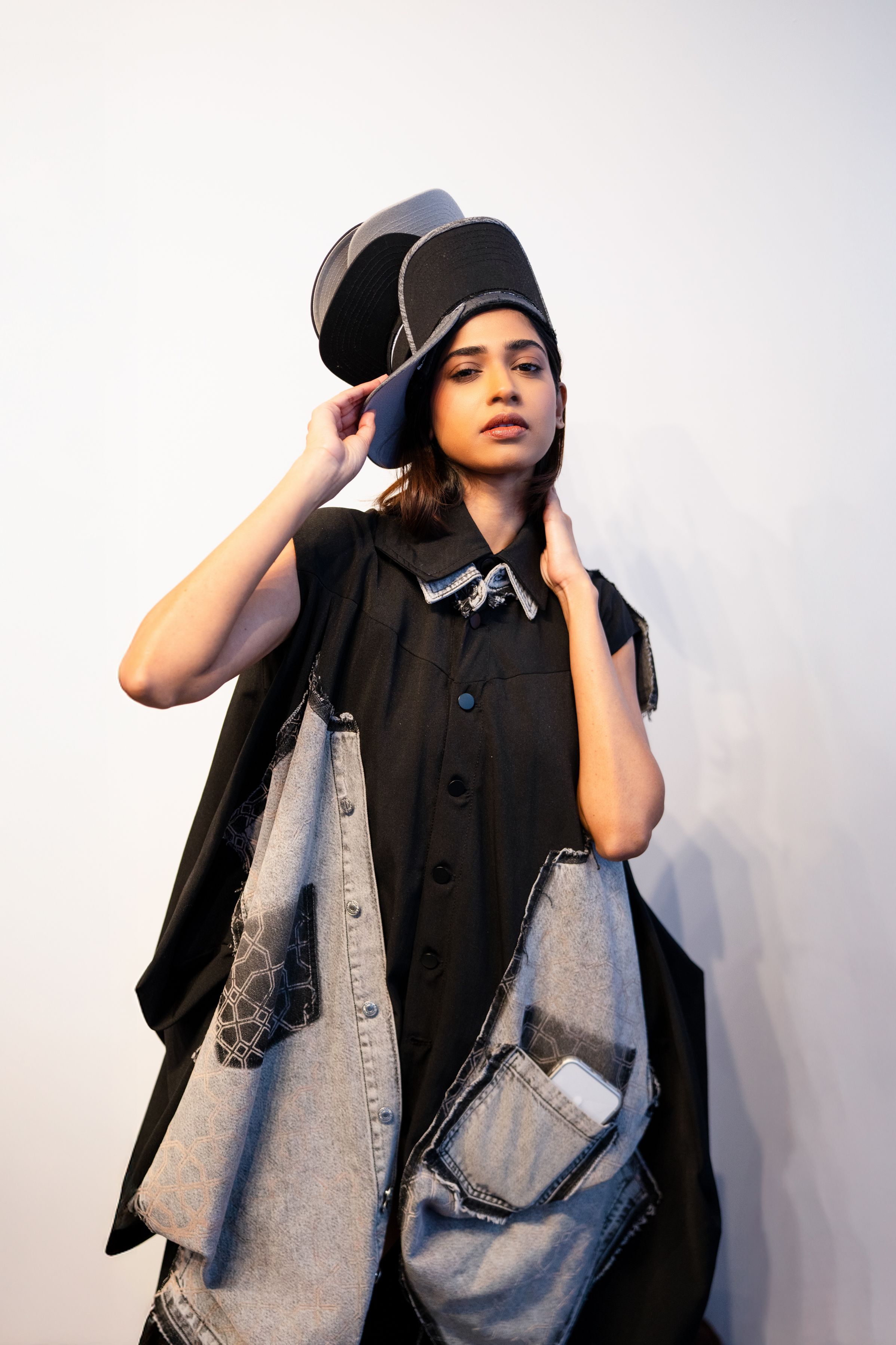 Woman in black oversized shirt and denim vest with large pockets, wearing a stylish black cap, posing against white background.
